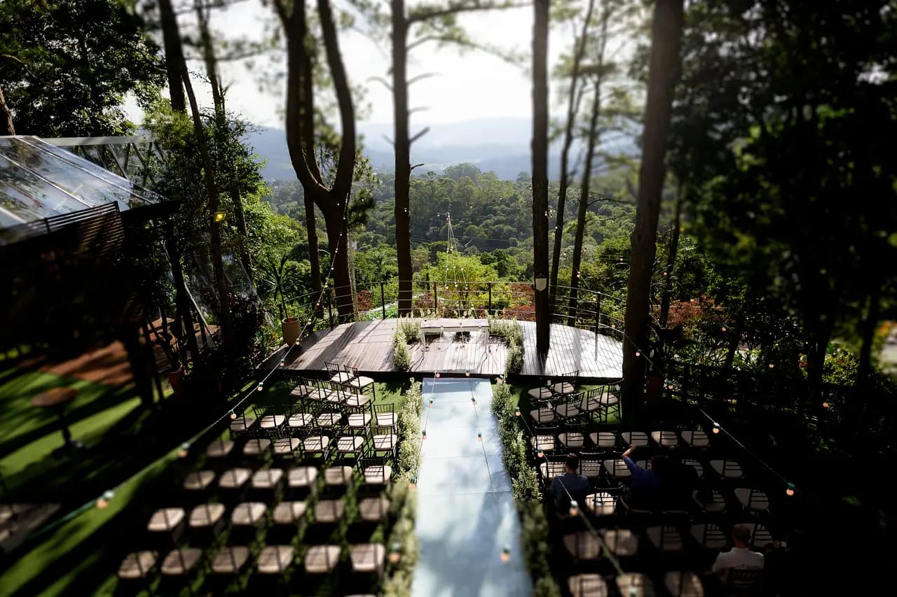Vista de cima da cerimônia na Villa Vezzane: o deck do altar entre as árvores, o corredor de vidro e as cadeiras dos convidados, com as montanhas ao fundo